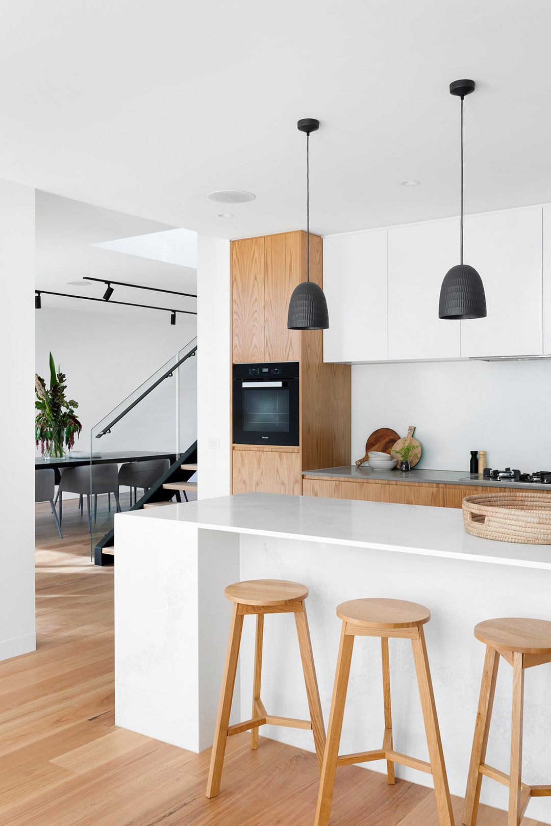 Kitchen with white cupboards, light wood accents and flooring; long white island with wooden bar stools
